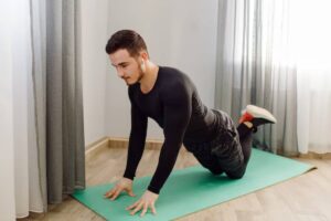 A person in a black long-sleeve shirt performs an exercise on a green yoga mat in a well-lit room with gray curtains.