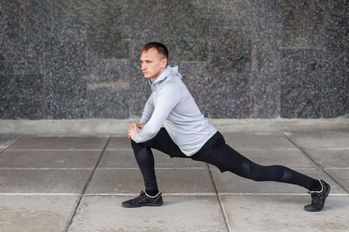 A person in workout gear performs a deep lunge on a stone pavement against a textured wall backdrop.
