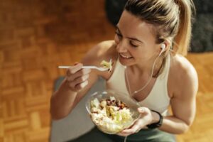 A person holding a bowl of salad with vegetables, listening to music through earphones in a cozy, wood-paneled setting.
