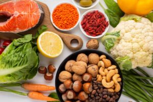 A colorful assortment of fresh vegetables, salmon, nuts, and seeds arranged on a white surface, showcasing healthy foods.