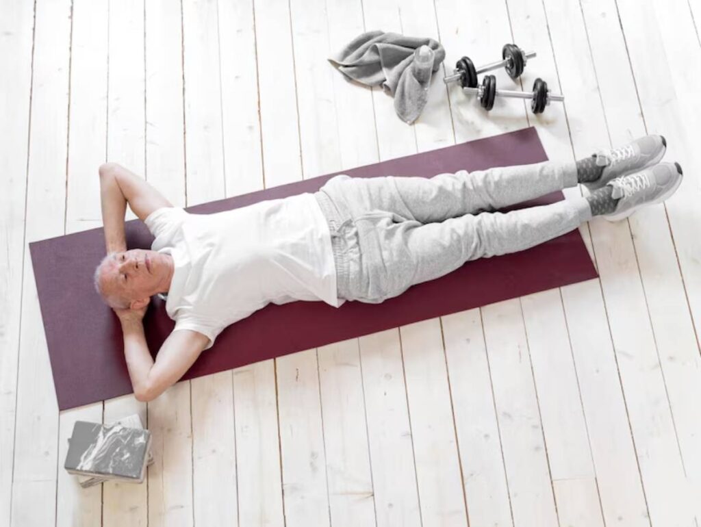An elderly person lies on a burgundy yoga mat, wearing casual workout clothes, with dumbbells and a towel beside them on a wooden floor.