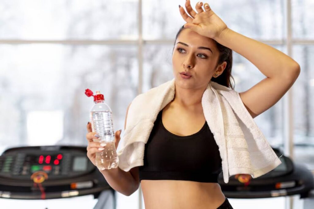 A fit person in a black tank top holds a water bottle and wipes sweat with a towel, standing near treadmills in a gym.