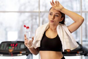 A fit person in a black tank top holds a water bottle and wipes sweat with a towel, standing near treadmills in a gym.