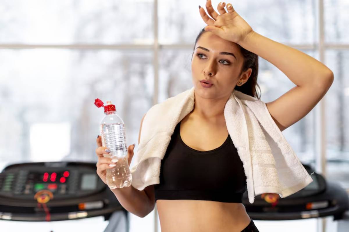 A fit person in a black tank top holds a water bottle and wipes sweat with a towel, standing near treadmills in a gym.