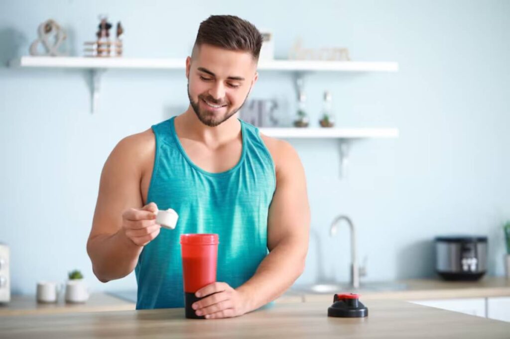 A man in a teal tank top holds a scoop above a red shaker bottle on a kitchen countertop, preparing a protein shake.