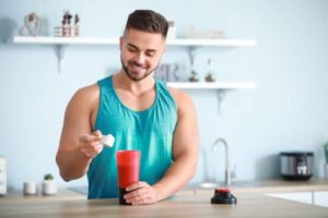 A man in a teal tank top holds a scoop above a red shaker bottle on a kitchen countertop, preparing a protein shake.