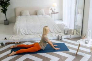 A person practices yoga on a blue mat in a cozy, minimalistic room with a bed, candles, and a leafy plant nearby.