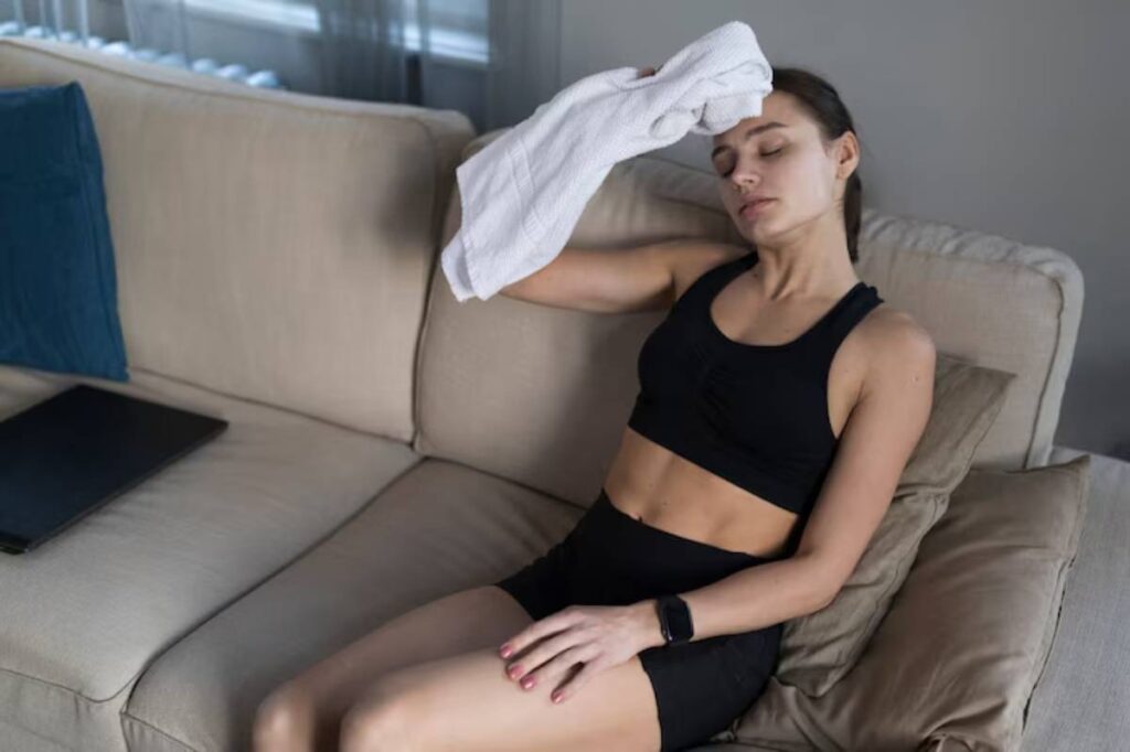 A person in black athletic wear sits on a couch, wiping their forehead with a towel, looking relaxed post-workout. A laptop rests nearby.