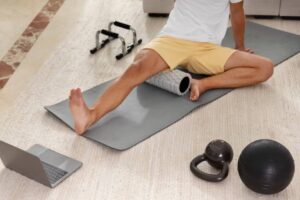 A person using a foam roller on a yoga mat with workout equipment and a laptop nearby in a home gym setting.