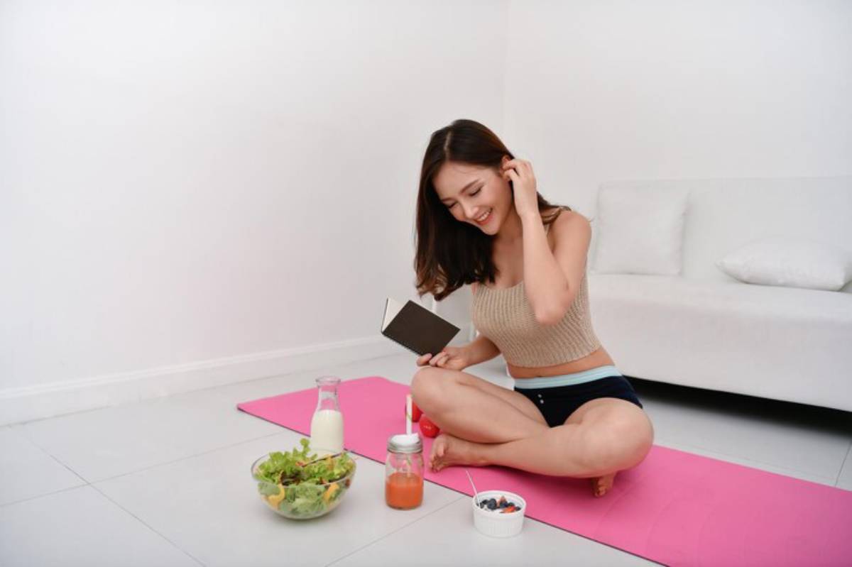 A woman sits cross-legged on a pink yoga mat, surrounded by healthy foods and a book in a bright, minimalistic room.