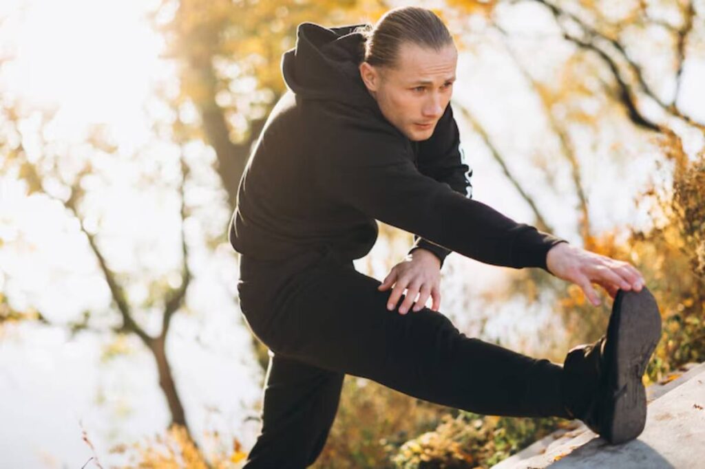 A person in a black hoodie stretches their leg while standing on a rocky surface surrounded by autumn foliage and soft sunlight.