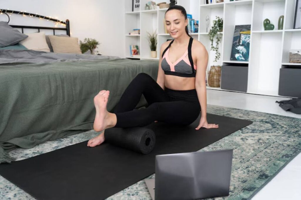 A person seated on a yoga mat uses a foam roller, with a laptop in front, in a cozy room with a bed and decorative shelves.