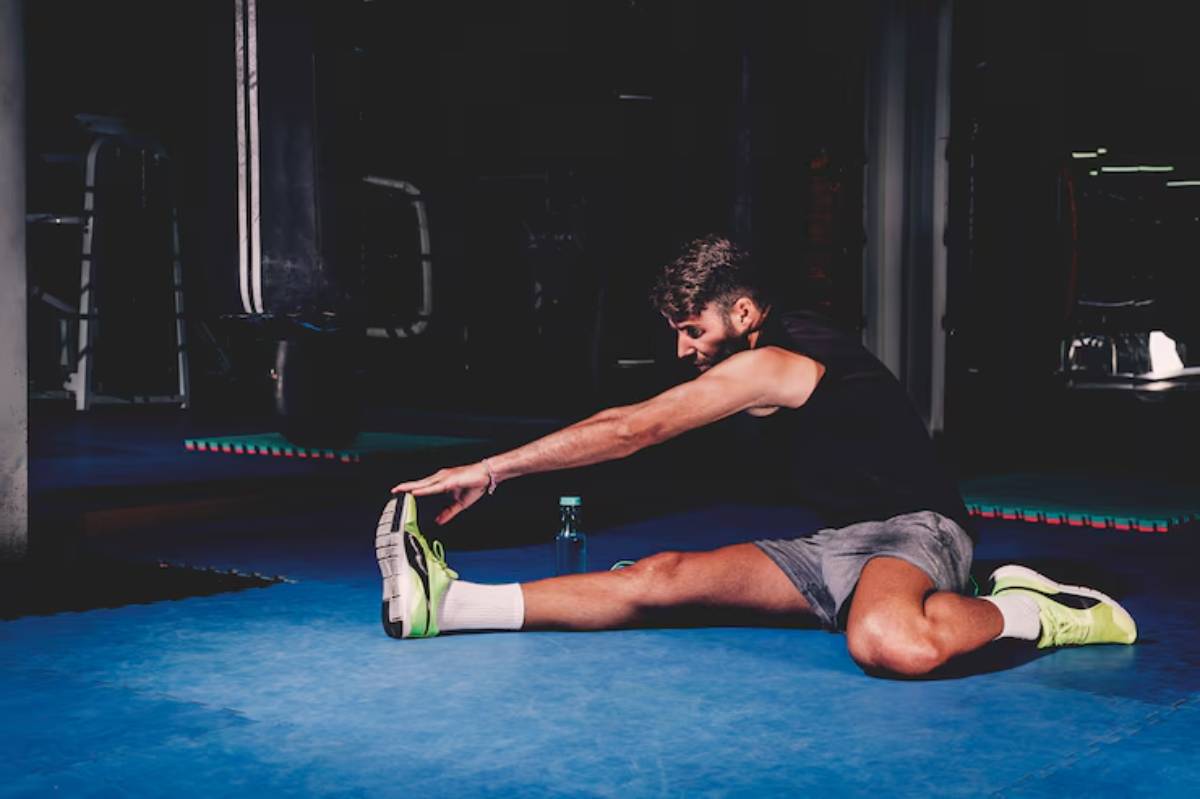 A person stretches their leg on a blue gym mat, wearing a black shirt and gray shorts, with a water bottle nearby.