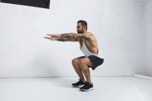 A man in a tank top and shorts performs a squat, demonstrating proper form in a minimalist gym setting.
