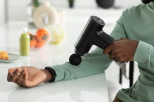 A person using a black massage gun on their arm while seated at a table with healthy snacks and drinks nearby.