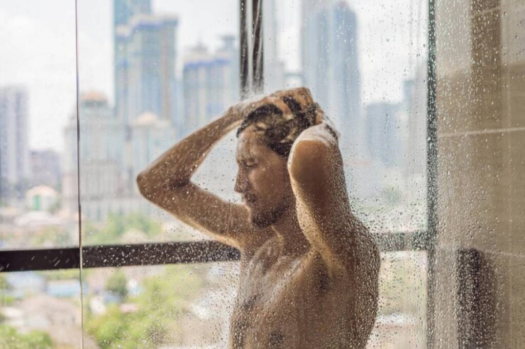 A person showers in a glass enclosure, with water streaming down and a city skyline visible in the background.