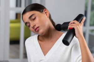 A person in a white sweater uses a black massage gun on their neck in a bright, modern living room setting.